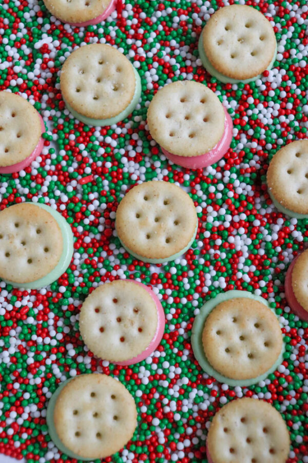 Round sandwich crackers with green and pink filling are scattered on a surface covered in red, green, and white nonpareil sprinkles.