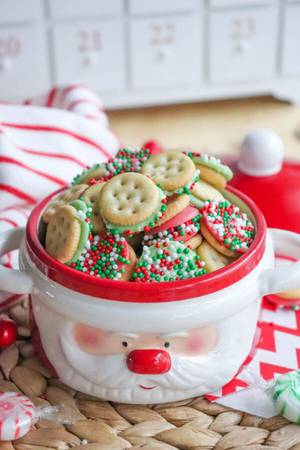 A festive Santa-themed bowl filled with round sandwich cookies decorated with red, white, and green sprinkles, sitting on a woven placemat with holiday decor in the background.