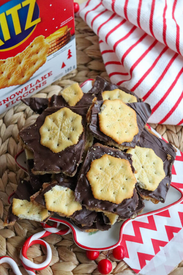 A plate of chocolate-covered Ritz crackers with snowflake patterns is displayed on a festive placemat, with a Ritz cracker box and a red-and-white striped cloth in the background.
