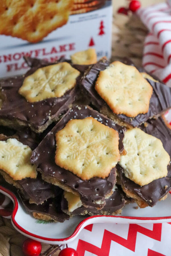 A plate filled with chocolate toffee bark topped with snowflake-shaped crackers, with a festive red and white napkin and holiday decorations in the background.
