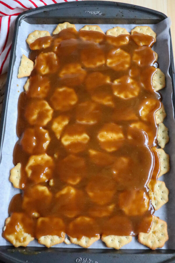 A baking tray lined with parchment paper holds a layer of hexagonal crackers covered in a thick, glossy caramel sauce. A red and white striped cloth is partially visible in the corner.