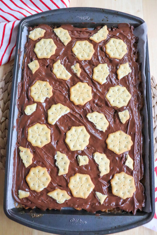 A baking tray lined with parchment paper holds a dessert topped with a layer of melted chocolate and scattered hexagonal crackers on top. A red-striped towel is partially visible in the background.