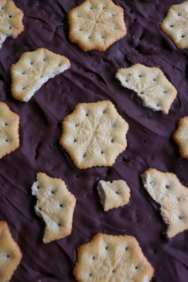 Hexagon-shaped cookies, some whole and some broken, are scattered on a dark, chocolate-covered surface. The cookies have a snowflake-like pattern stamped on top.