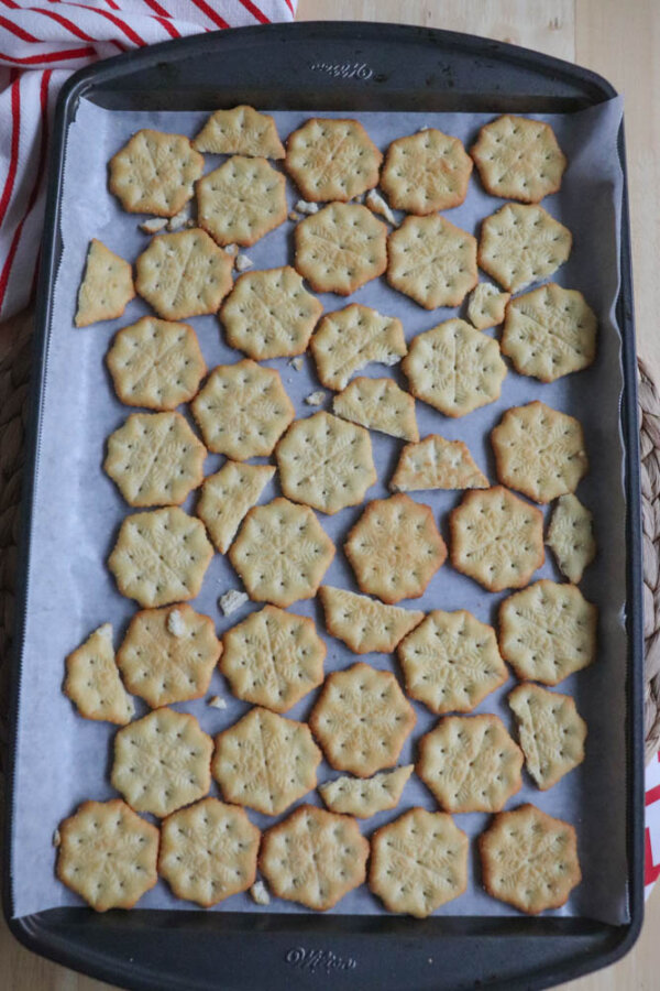 A baking tray lined with parchment paper holds several golden-brown, hexagonal crackers with a decorative pattern. Some crackers are broken, and a red-and-white towel is partially visible in the upper left corner.