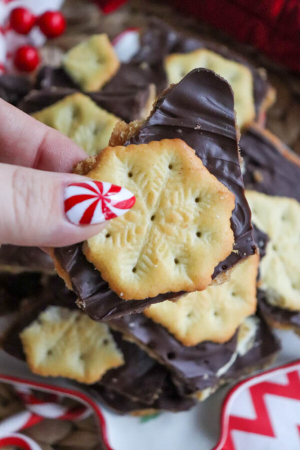 A hand with red and white peppermint swirl nail art holds a piece of chocolate-coated cracker bark topped with a snowflake-patterned cracker. More pieces are stacked in the background with festive red accents nearby.