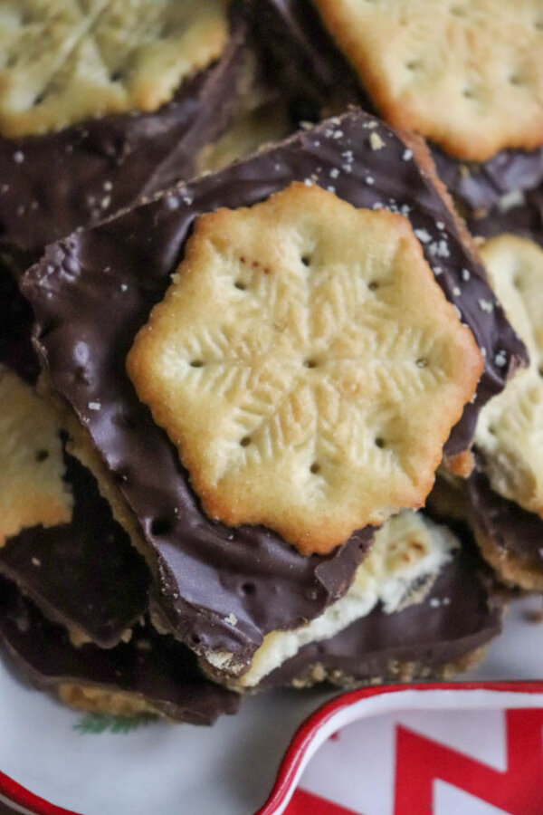 A close-up of square chocolate-covered crackers topped with round cookies featuring a snowflake design, stacked on a plate with a red and white chevron edge.