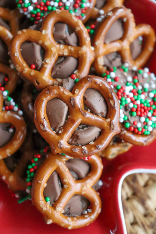 A close-up of pretzels topped with melted chocolate and colorful red, green, and white sprinkles, arranged on a red dish.