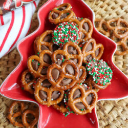 A red, tree-shaped dish filled with pretzel bites topped with melted Rolo candies; some are decorated with red, white, and green sprinkles. A Rolo candy roll sits nearby on a woven placemat.
