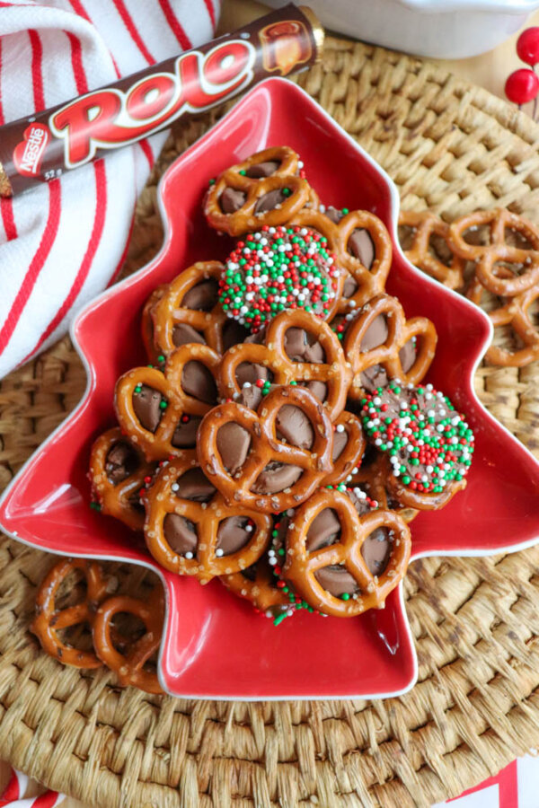 A red, tree-shaped dish filled with pretzel bites topped with melted Rolo candies; some are decorated with red, white, and green sprinkles. A Rolo candy roll sits nearby on a woven placemat.