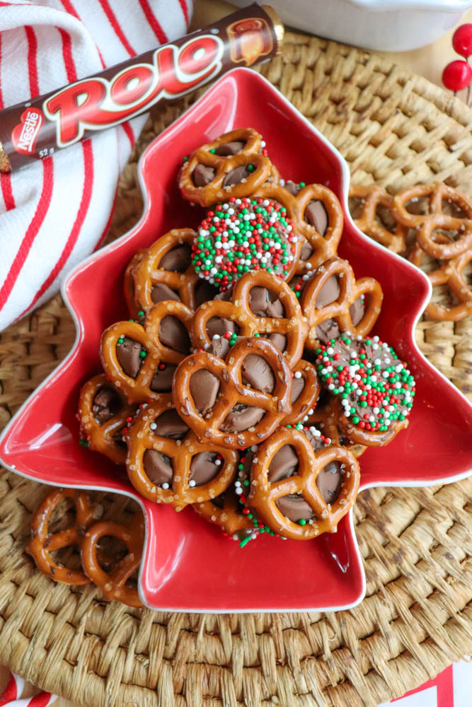 A red, tree-shaped dish filled with pretzel bites topped with melted Rolo candies; some are decorated with red, white, and green sprinkles. A Rolo candy roll sits nearby on a woven placemat.