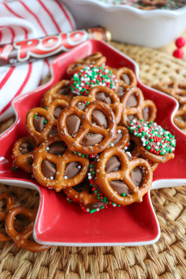A red, tree-shaped dish holds pretzels topped with melted chocolate candies, some decorated with red, white, and green sprinkles. A Rolo candy wrapper and more treats are visible in the background.