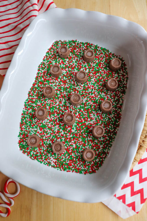 A white baking dish with a layer of red, green, and white sprinkles, topped with evenly spaced Rolo chocolates. Red-and-white napkins and ribbon are visible beside the dish on a wooden surface.