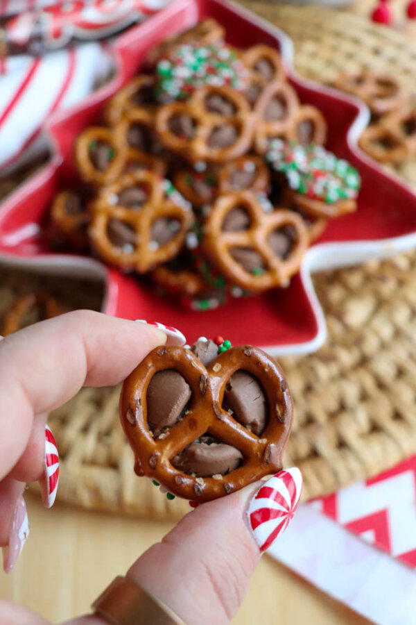 A hand with holiday-themed nails holds a chocolate-filled pretzel in front of a tree-shaped plate of more chocolate pretzels decorated with festive sprinkles.