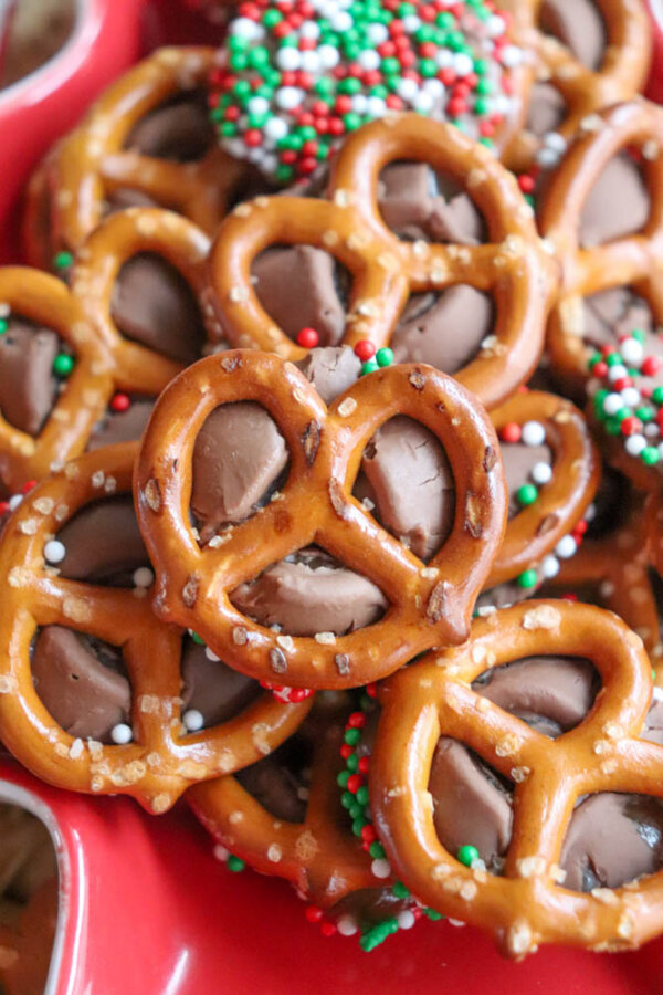 A close-up of pretzels topped with melted chocolate and festive red, green, and white sprinkles, arranged on a red platter.