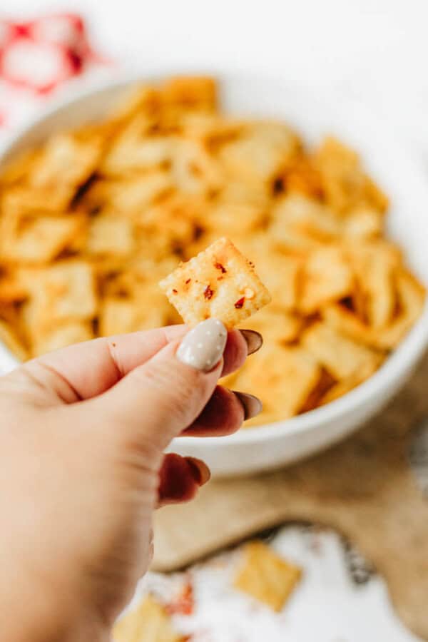 A hand with manicured nails holds a square, orange-colored cracker with visible seasoning in front of a bowl filled with similar crackers. The background is softly blurred.