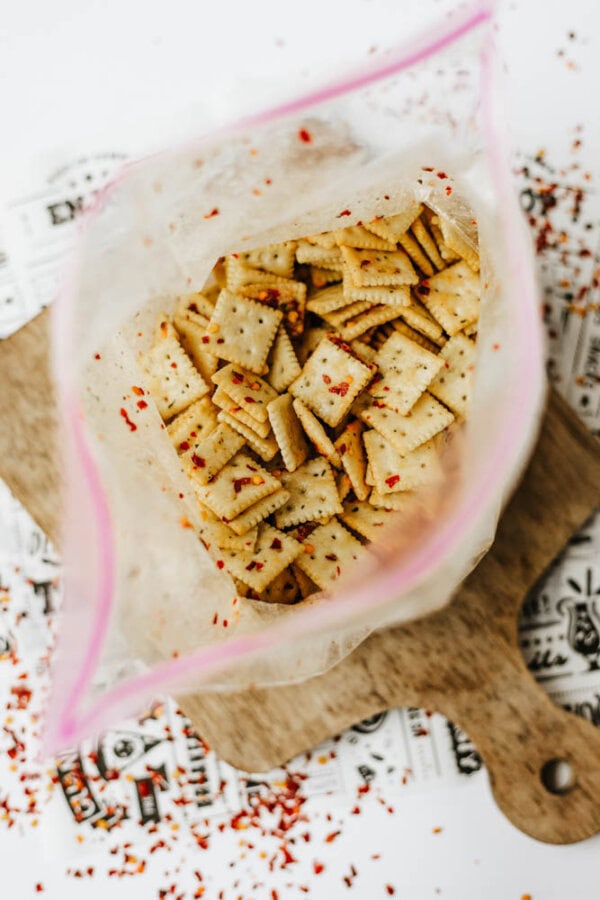An open plastic bag filled with square crackers sprinkled with red chili flakes, placed on a wooden cutting board with some crackers and chili flakes scattered around.