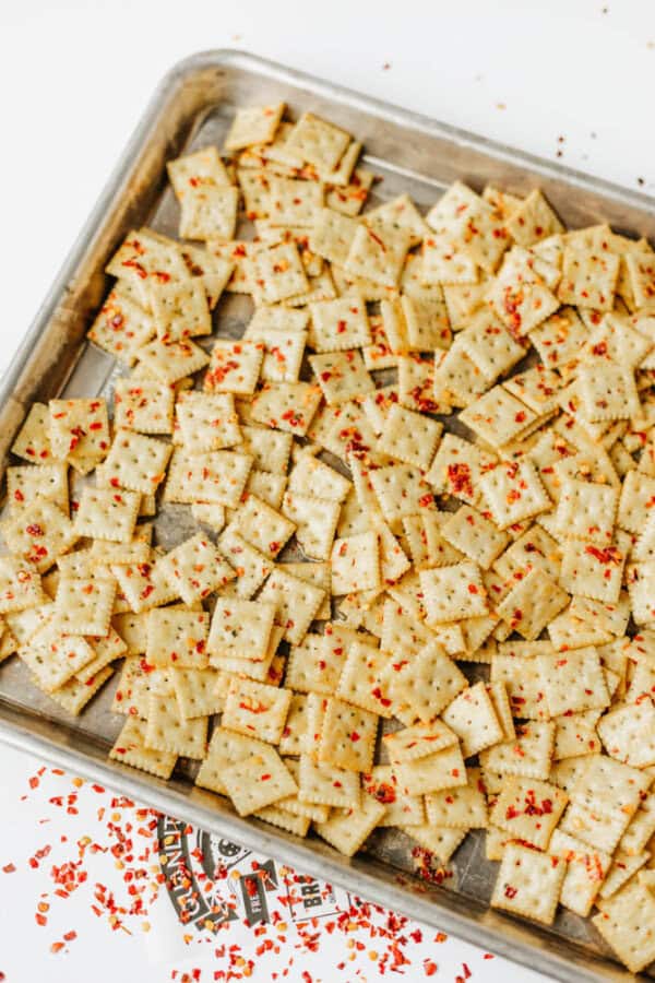A metal baking sheet filled with square crackers sprinkled with red seasoning flakes, some seasoning scattered on the white surface nearby.
