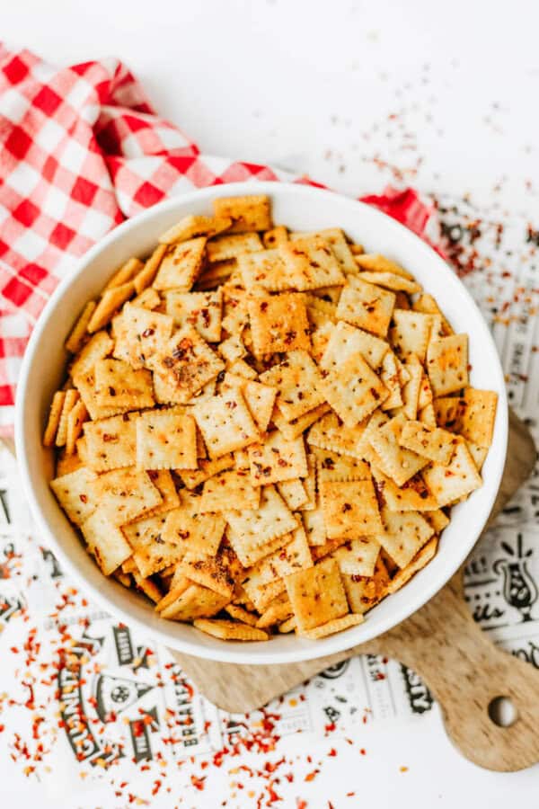 A white bowl filled with seasoned square crackers sits on a patterned paper and wooden board, next to a red and white checkered cloth. Cracker crumbs and seasoning are scattered around.
