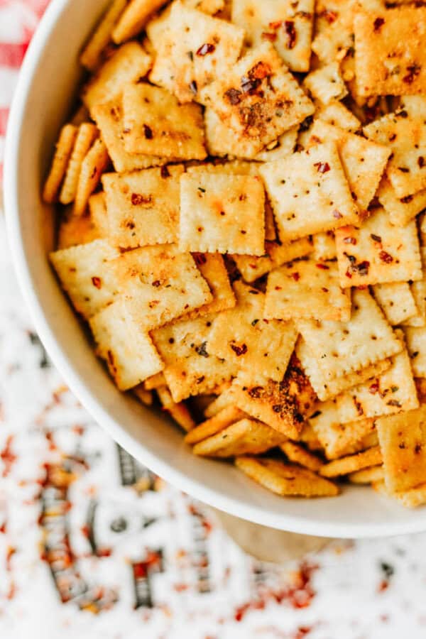 A bowl filled with square, seasoned crackers topped with red pepper flakes and spices, sitting on a patterned surface.