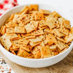 A white bowl filled with seasoned square crackers sits on a wooden board, with a red-patterned cloth and decorative paper in the background.
