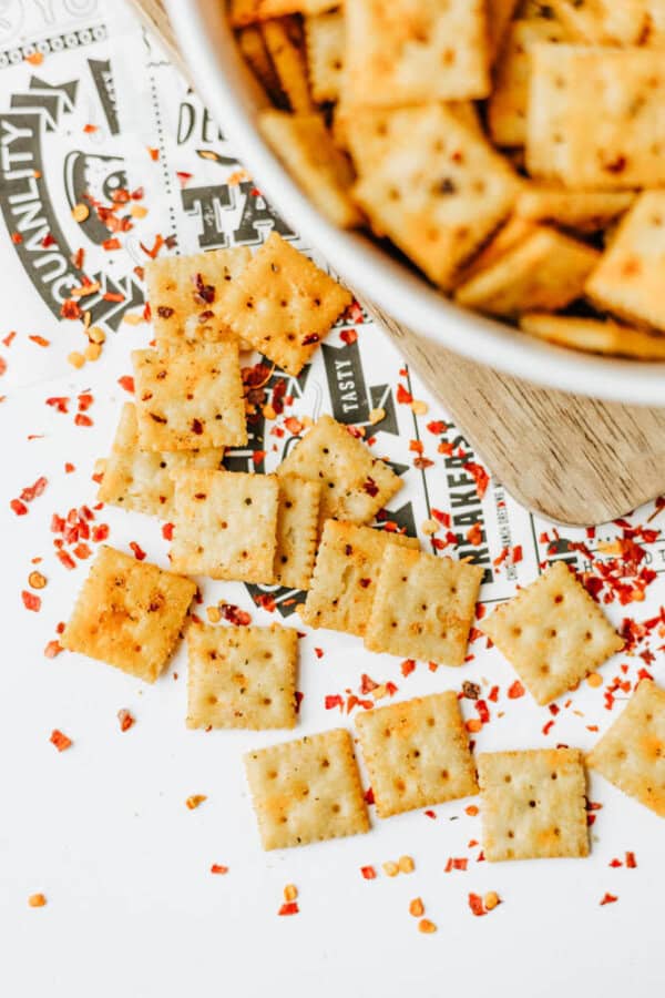 Square crackers sprinkled with red pepper flakes are scattered on a white surface with printed paper underneath. A bowl filled with more crackers is partially visible in the top right corner.