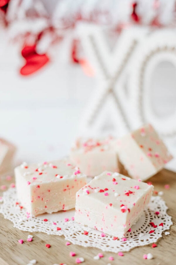 Four pieces of white fudge with red and pink sprinkles rest on a lace doily atop a wooden surface, with a blurred background featuring red decorations and a decorative XO sign.