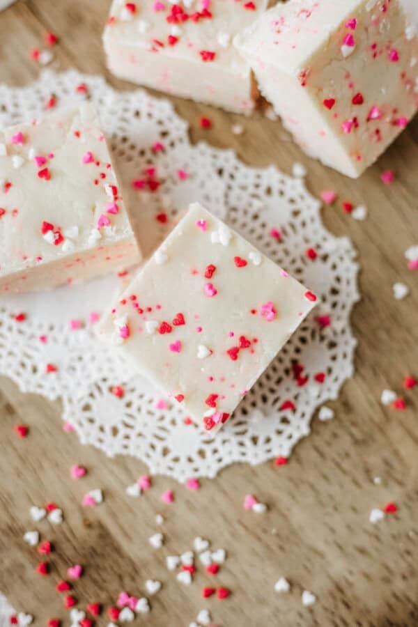 Squares of white fudge with red, pink, and white heart-shaped sprinkles rest on a white paper doily atop a wooden surface, with sprinkles scattered around.