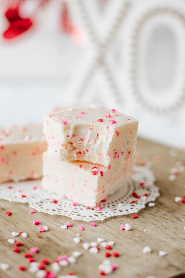 Two pieces of white fudge with pink and red heart sprinkles stacked on a wooden surface, one with a bite taken out, placed on a lace doily. Sprinkles are scattered around, with white decor in the background.