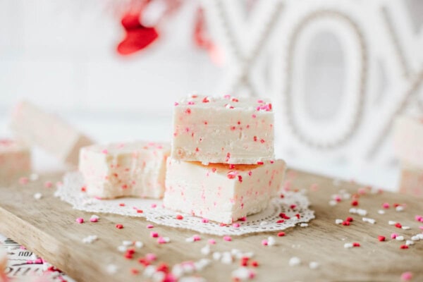 Three pieces of white fudge with red and pink sprinkles are stacked on a wooden board with a lace doily, with more fudge pieces and Valentine’s-themed decorations blurred in the background.