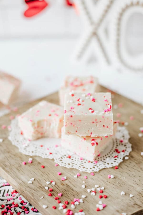 White fudge squares topped with pink, red, and white heart-shaped sprinkles are stacked on a wooden board with a lace doily, surrounded by more sprinkles. Valentines Day decorations are slightly blurred in the background.