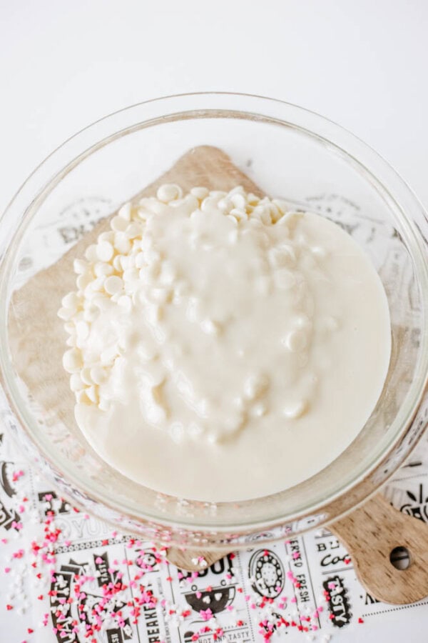 A glass bowl filled with white chocolate chips and sweetened condensed milk sits on a wooden board, surrounded by pink and red sprinkles on a decorative paper.