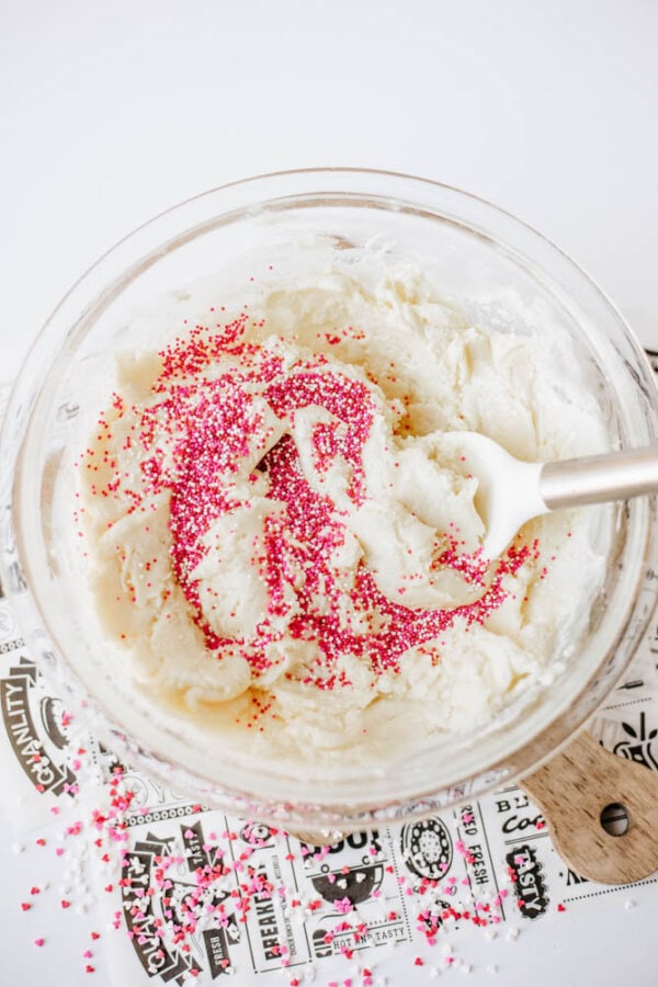 A glass bowl filled with cookie dough and pink sprinkles being mixed with a white spatula, set on a patterned paper and wooden cutting board. Some sprinkles are scattered around the bowl.