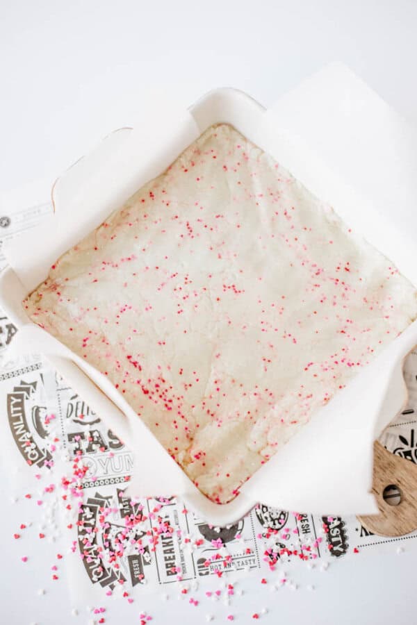 A square baking pan lined with parchment paper holds unbaked dough topped with pink and red sprinkles. The pan sits on top of black and white decorative papers, with more sprinkles scattered nearby.