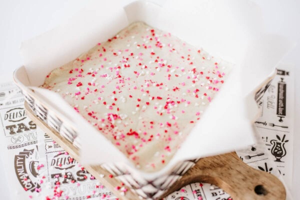 A square baking pan lined with parchment paper holds unbaked batter topped with red, pink, and white heart-shaped sprinkles. The pan rests on a wooden board and decorative paper.