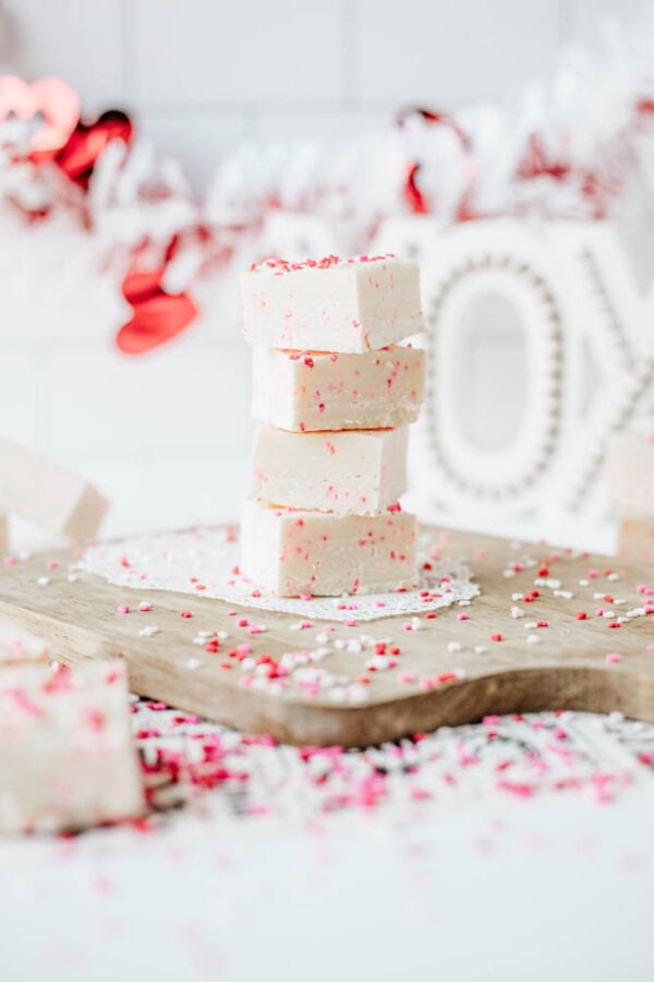 A stack of white fudge squares topped with red and white sprinkles sits on a wooden board, surrounded by more fudge pieces and sprinkles. Valentines Day-themed decorations appear blurred in the background.