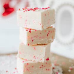 Four pieces of white fudge with red and pink sprinkles are stacked on a lace doily, with more sprinkles scattered around on a light surface. The background is softly blurred.