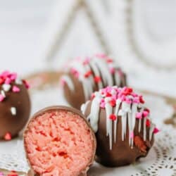 Valentine’s Day cake balls coated in chocolate and decorated with pink, red, and white sprinkles and icing, displayed on a doily-covered wooden surface. One cake ball is cut open to show the pink interior.