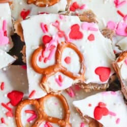 Valentines Day pretzel toffee topped with white chocolate, pink and red heart sprinkles, and whole pretzels, arranged on a surface with a homemadeheather.com label at the bottom.