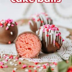 Chocolate-covered strawberry cake balls, some decorated with pink, red, and white sprinkles, sit on a doily. One cake ball is cut open, showing a pink interior. Text reads strawberry cake balls and SO GOOD.