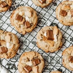 A cooling rack with s’mores cookies topped with chocolate pieces and mini marshmallows, placed on newspaper with extra marshmallows nearby. Text reads smores cookies and homemadeheather.com.