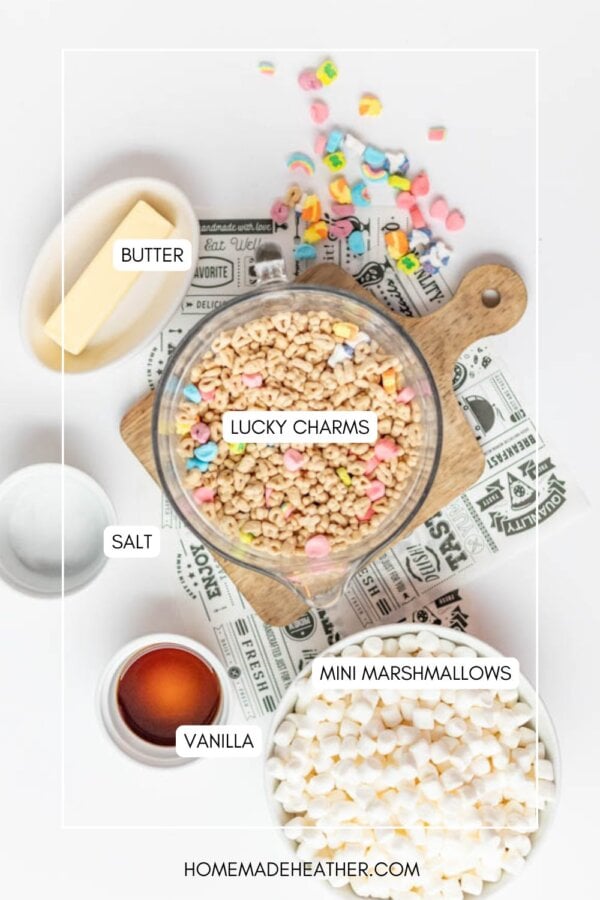 Top-down view of ingredients for a recipe, including a bowl of Lucky Charms cereal, a plate of butter, a dish of salt, a bowl of mini marshmallows, and a small bowl of vanilla extract on a white surface.