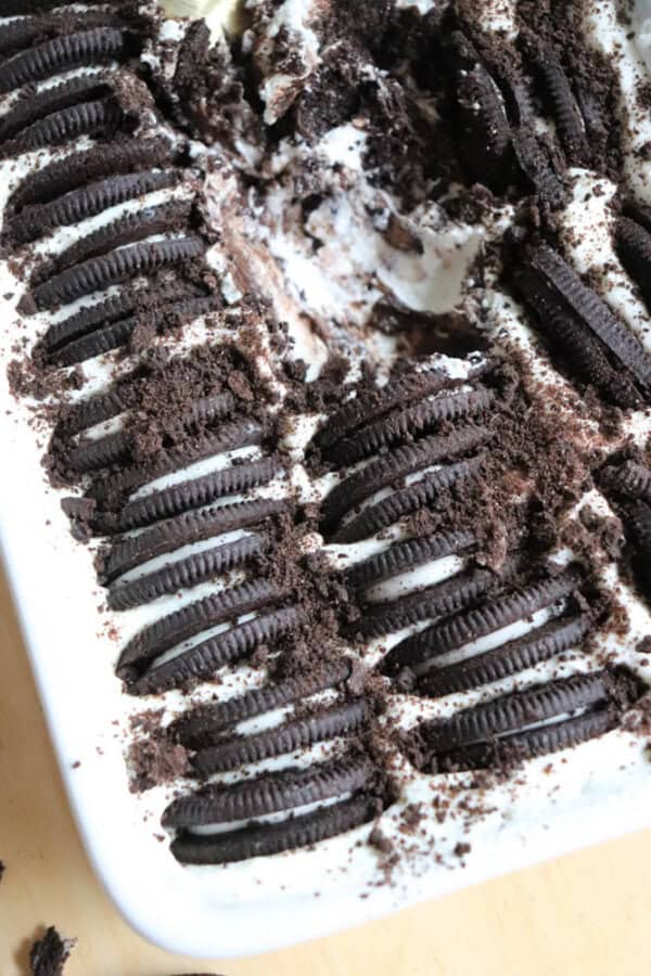 A close-up of a dessert topped with rows of whole chocolate sandwich cookies and crumbled cookies over a layer of whipped cream or frosting in a rectangular dish. Some portions have been scooped out.