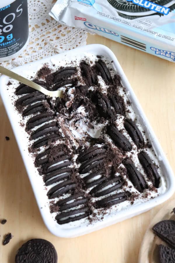 A rectangular dish filled with a layered dessert topped with whole and crushed Oreo cookies. A spoon is partially scooping out a serving. An open package of gluten-free Oreos and a drink are nearby on the table.