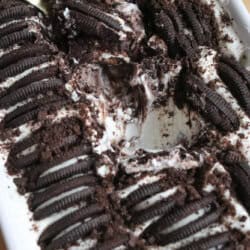 A rectangular dish filled with cookies and cream ice cream, topped with rows of chocolate sandwich cookies and cookie crumbs, with a section scooped out and a metal serving spoon resting inside.