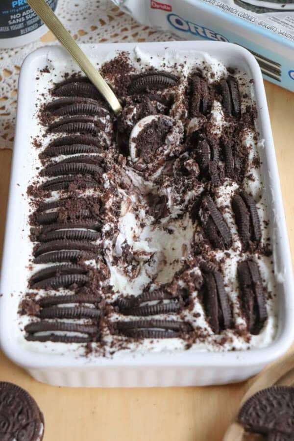 A rectangular dish filled with creamy dessert topped with rows of Oreo cookies and cookie crumbs, with a spoon scooping out a portion from the center.