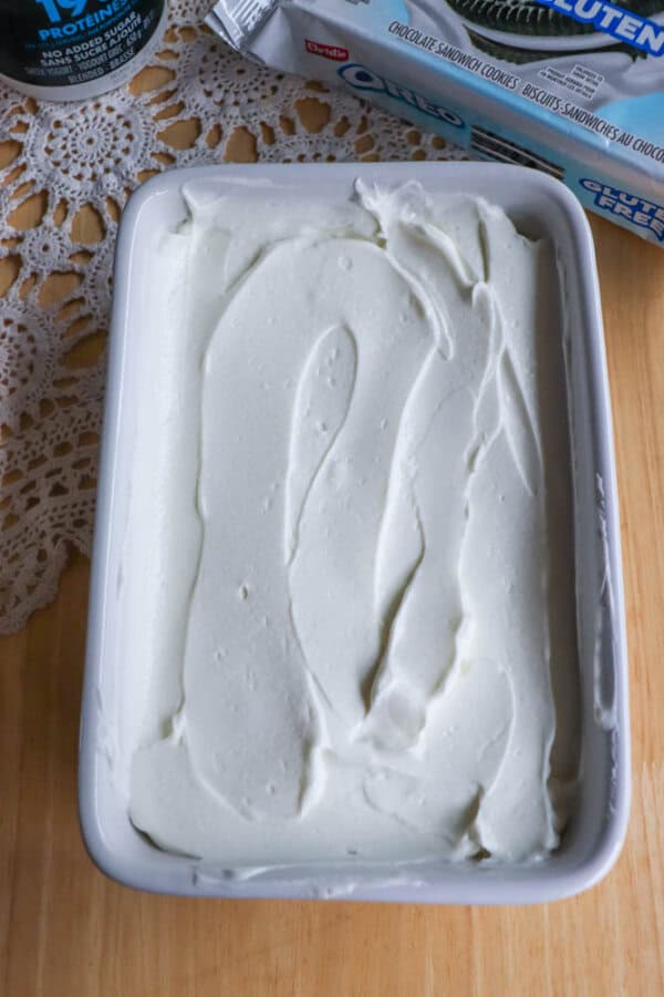 A rectangular white dish filled with smooth, creamy white frosting or whipped mixture sits on a wooden surface near a lace doily and partially visible packaging.
