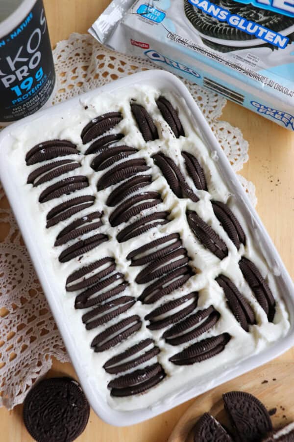 A white baking dish filled with a creamy dessert topped with rows of halved gluten-free Oreo cookies. A container of yogurt and a package of gluten-free Oreos are nearby on a wooden table with a doily.