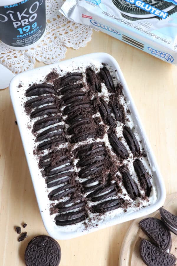 A rectangular dish of cookies and cream dessert topped with whole and crushed chocolate sandwich cookies sits on a wooden table, with a doily, yogurt cup, and cookie packages nearby.