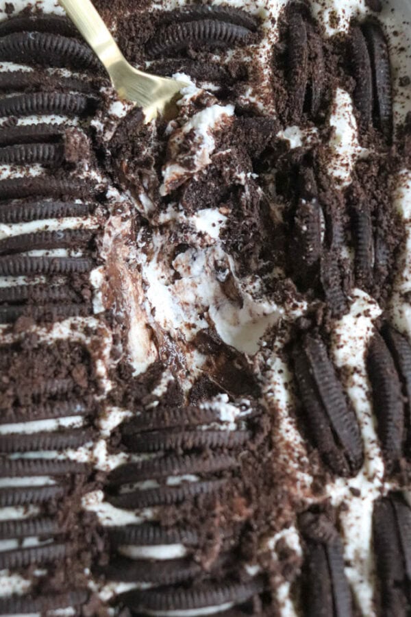 A close-up of a layered dessert with whole Oreo cookies, whipped cream, and chocolate, partially scooped with a spoon. Crumbs and cream are visible in the textured layers.