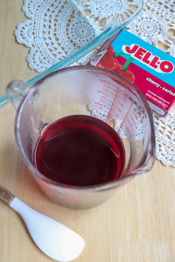 A measuring cup filled with red liquid sits on a wooden table next to a cherry Jello box, a white spatula, and a decorative lace doily.